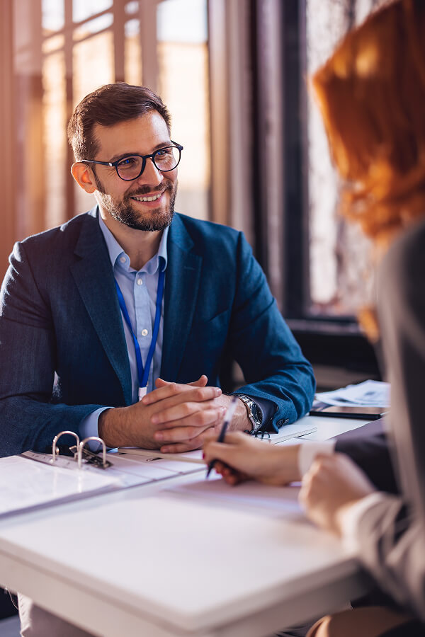 Smiling man in a business suit having a professional conversation across a desk with a woman taking notes.