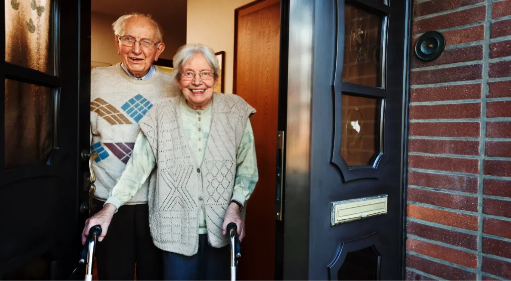 Elderly couple standing together in their front doorway, with the woman using a walker, representing adults who may need conservatorship protection in California.