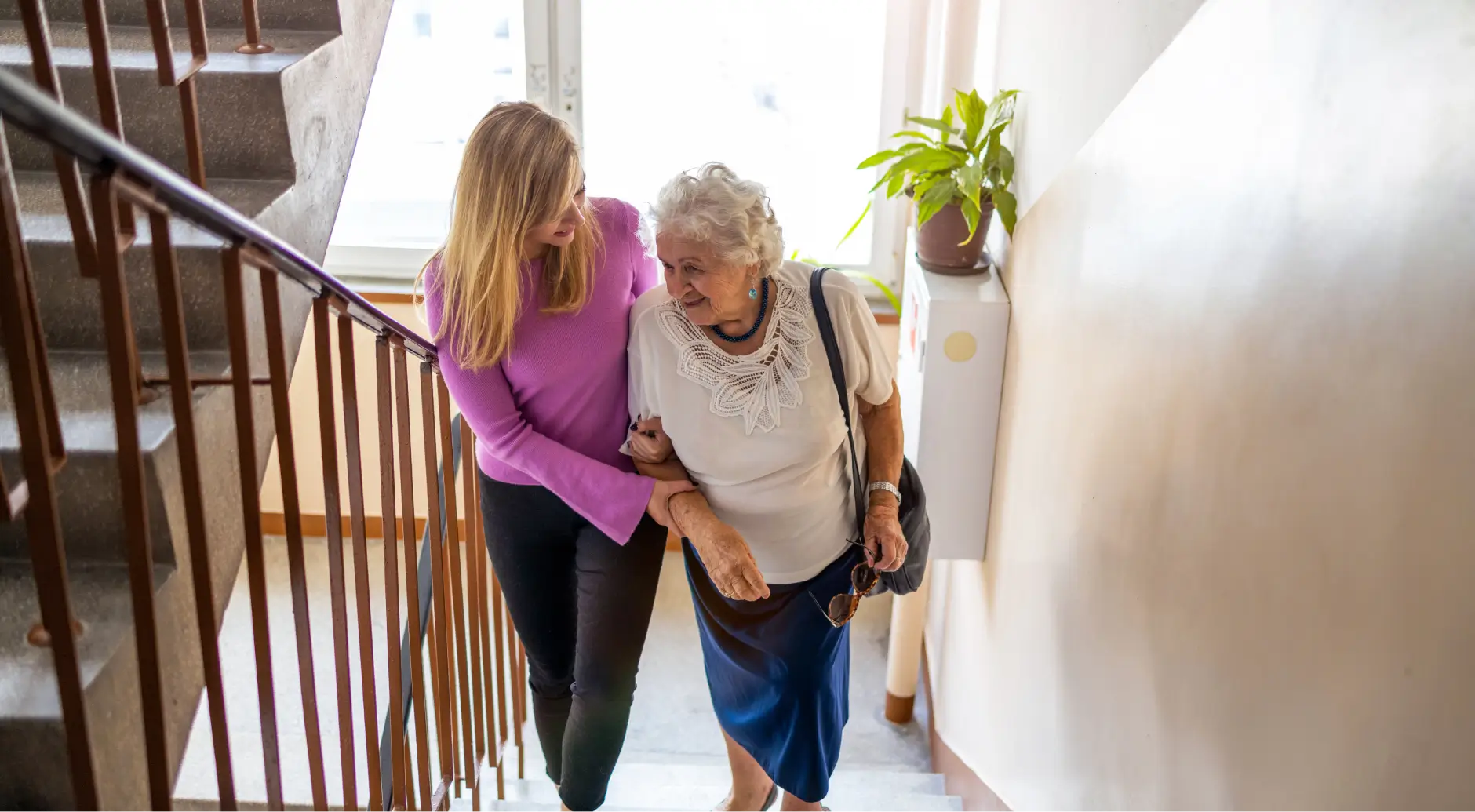 Younger woman helping an elderly woman walk up a staircase in a residential building, illustrating the caregiving role central to conservatorship and guardianship in California.