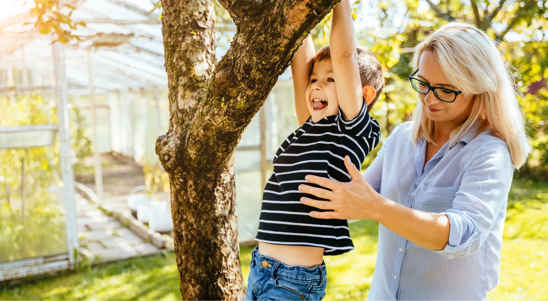 Young boy laughing while hanging from a tree branch as a woman supports him in a sunny backyard, representing the guardian-child relationship in California guardianship cases.